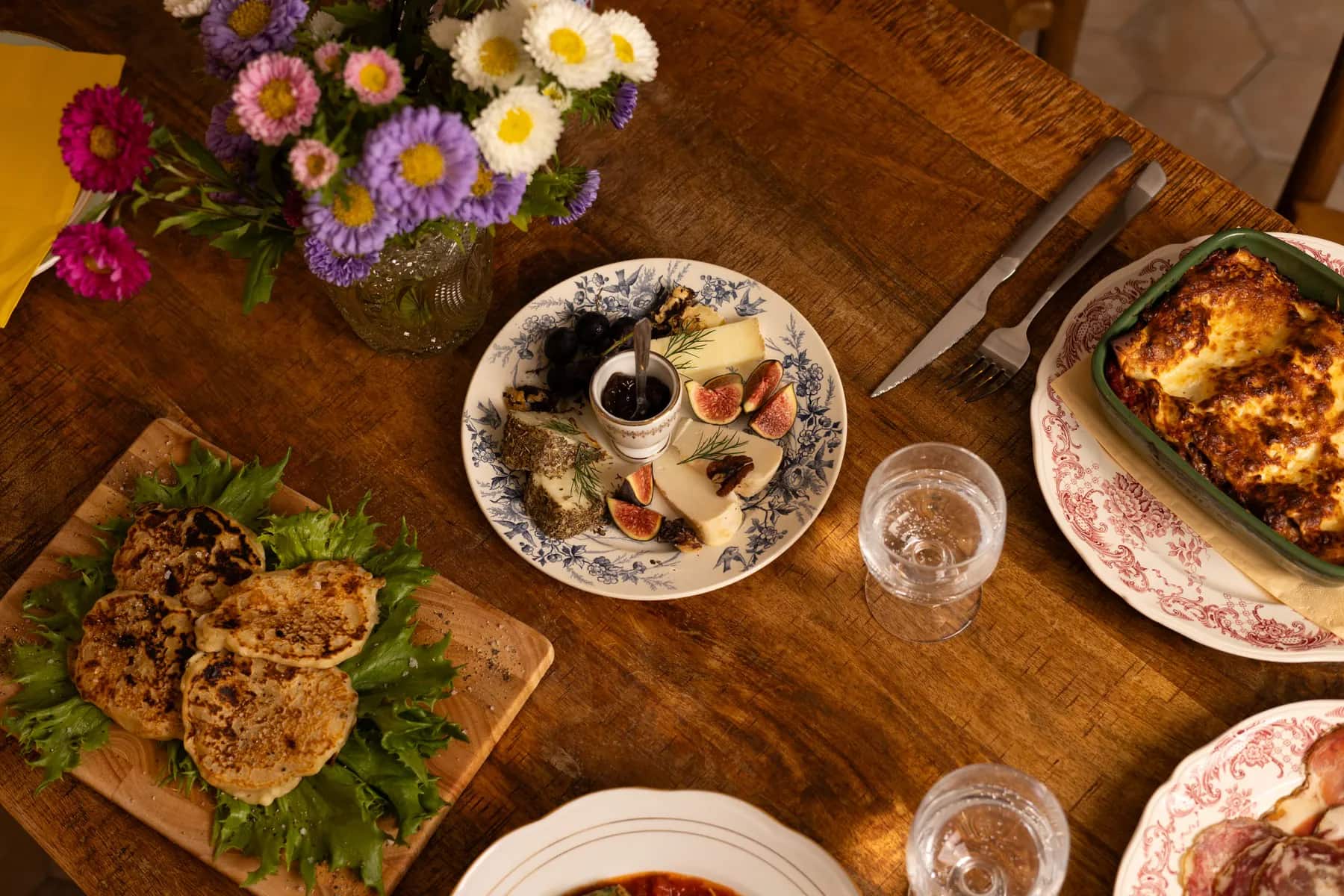 Une table garnie vue d’en haut : galettes, assiette de fromages, gratin et bouquet de fleurs.