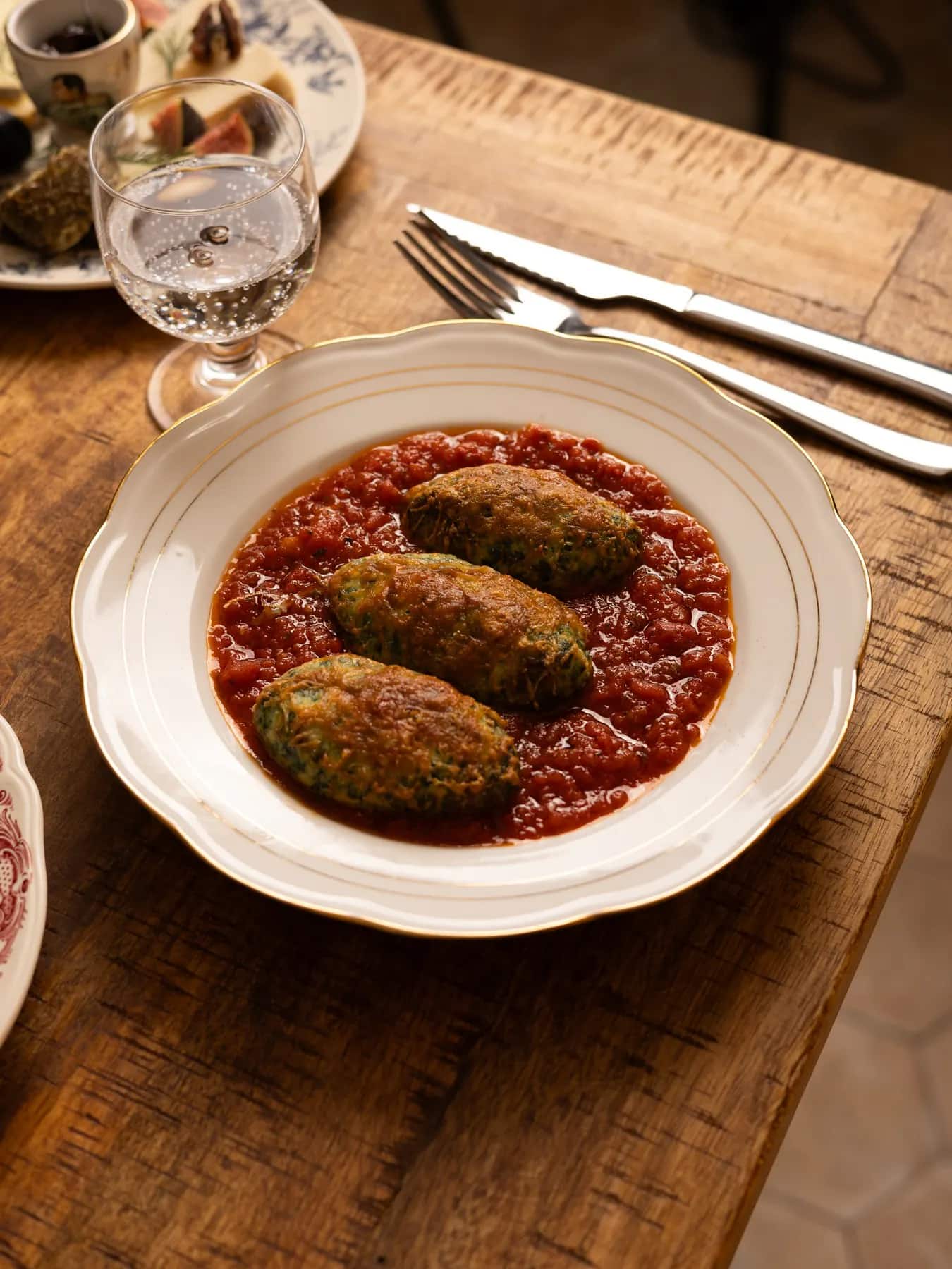 Storzapretti : quenelles de brocciu gratinées à la tomme de brebis, sur une sauce tomate aux herbes du maquis, dans une assiette à liseré doré.