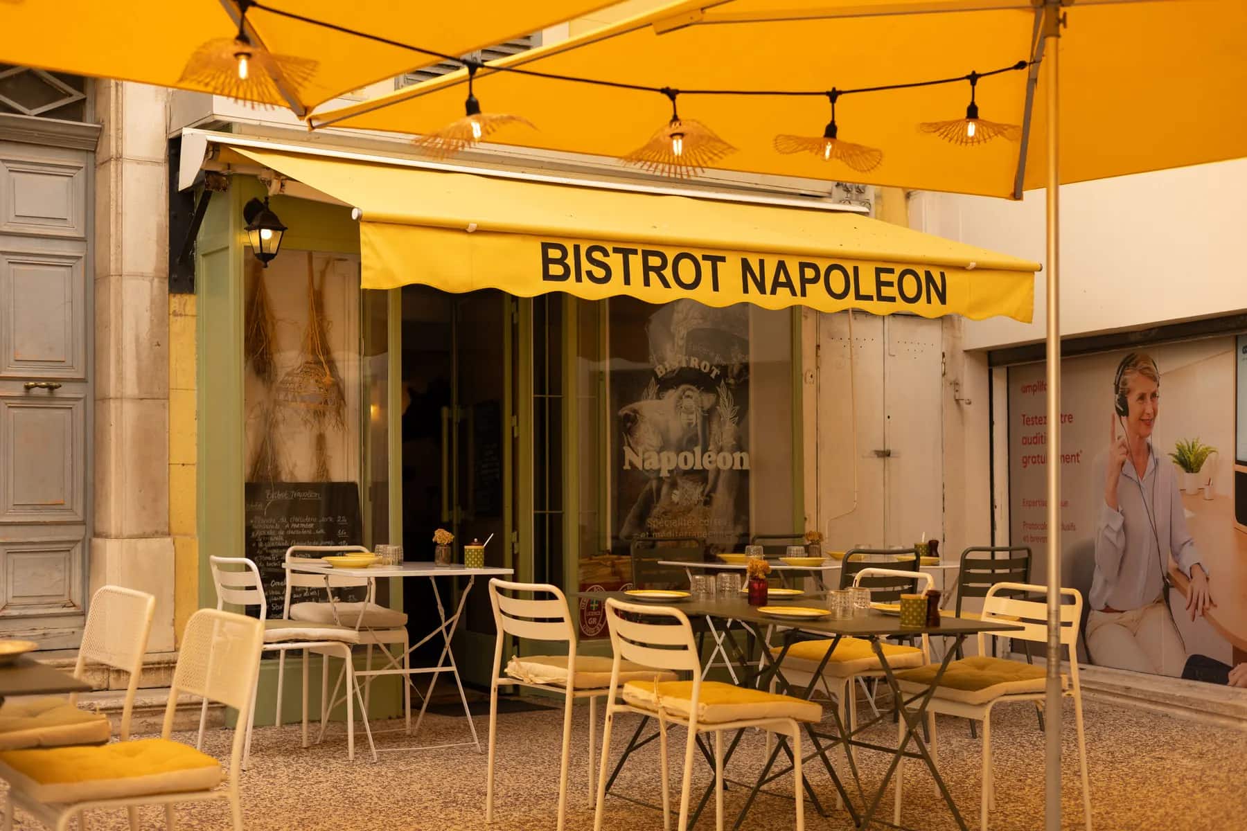 La terrasse sous la marquise jaune « Bistrot Napoléon », guirlandes lumineuses et chaises bistrot.